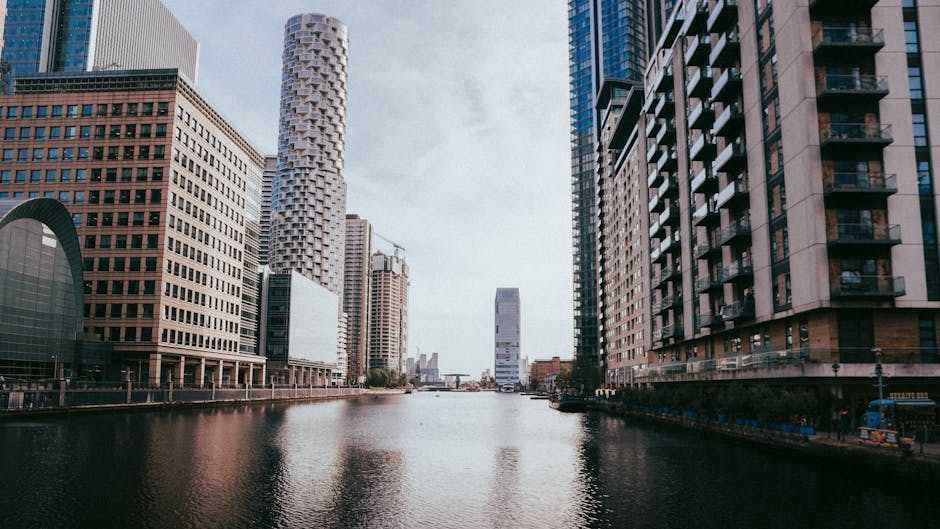 A daytime view of Canary Wharf in London featuring a wide river canal with calm water reflecting the surrounding high-rise buildings. On the left side, modern office towers with glass facades and geometric architectural designs are visible, with some buildings under construction, indicated by cranes. To the right, there is a tall residential building with balconies and a beige exterior. In the distance, a sleek, white, curved skyscraper stands prominently against a partly cloudy sky. The scene captures the urban environment typical of Docklands, illustrating a typical waterfront skyline. This image relates to house removals and moving services, such as those offered by Docklands Man and Van, highlighting the importance of careful planning when transporting furniture and belongings through high-rise environments. The setting suggests an area where moving specialists may need to navigate high-rise access, loading zones, and the use of transport equipment, including vans and trolleys, for efficient home relocation and furniture transport.