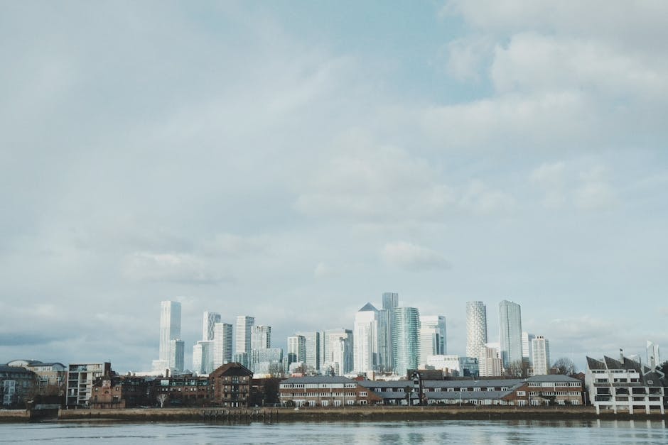 A view of Canary Wharf's high-rise office buildings and modern skyscrapers in the London Docklands area, with a partly cloudy sky overhead. In the foreground, a row of low-rise brick and white residential buildings lines the riverbank. The scene is captured from across the water, emphasizing the contrast between the older residential structures and the new, glass-clad towers. Visible in the image are a few boats on the river, with some construction equipment and materials possibly seen near the waterfront, suggesting ongoing building or maintenance work. This setting provides a relevant context for house removals or furniture transport, highlighting the urban environment involved in high-rise moves or home relocation projects. The image is naturally suited for content about packing, loading, and logistics, with the Docklands Man and Van company mentioned subtly for reference to professional moving services in this district.