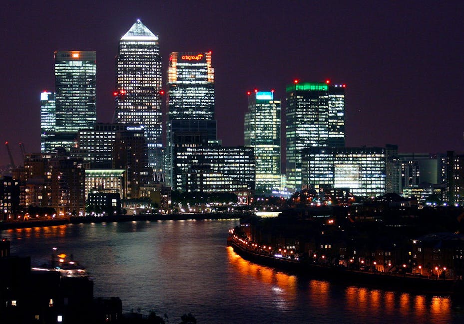 A daytime view of Canary Wharf in London featuring a wide river canal with calm water reflecting the surrounding high-rise buildings. On the left side, modern office towers with glass facades and geometric architectural designs are visible, with some buildings under construction, indicated by cranes. To the right, there is a tall residential building with balconies and a beige exterior. In the distance, a sleek, white, curved skyscraper stands prominently against a partly cloudy sky. The scene captures the urban environment typical of Docklands, illustrating a typical waterfront skyline. This image relates to house removals and moving services, such as those offered by Docklands Man and Van, highlighting the importance of careful planning when transporting furniture and belongings through high-rise environments. The setting suggests an area where moving specialists may need to navigate high-rise access, loading zones, and the use of transport equipment, including vans and trolleys, for efficient home relocation and furniture transport.