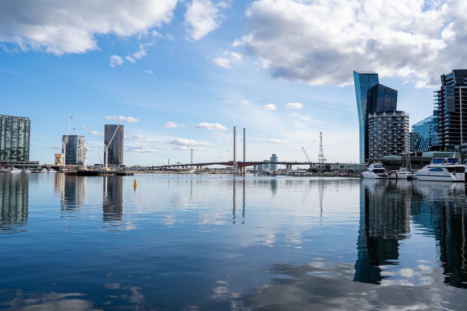 A wide view of a modern cityscape with several high-rise buildings and a distinctive curved glass skyscraper along the waterfront. The calm water in the foreground reflects the buildings and a partly 