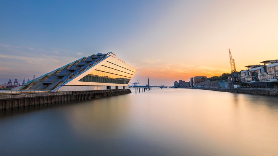 A wide view of a modern cityscape with several high-rise buildings and a distinctive curved glass skyscraper along the waterfront. The calm water in the foreground reflects the buildings and a partly 