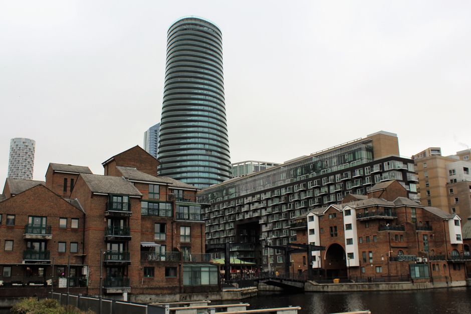 A panoramic view of a modern city skyline with numerous high-rise office buildings and residential towers made of glass and steel, situated behind a row of older, low-rise brick and wooden residential