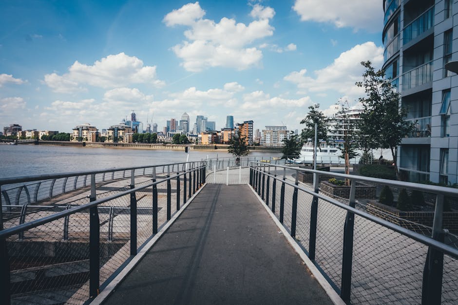 A wide, elevated walkway with metal railings and a non-slip surface extends over the edge of a riverfront in the Docklands area, offering a view of the city skyline with modern high-rise buildings and construction cranes in the distance under a partly cloudy sky. The walkway is adjacent to contemporary residential apartments on one side, with some small trees and outdoor seating areas visible along the path. This scene depicts the environment where a house or flat relocation might take place, with natural light illuminating the scene, suitable for illustrating house removals or furniture transport in an urban waterfront setting. Docklands Man and Van uses such locations to facilitate efficient loading and unloading processes during home relocation or moving services, highlighting the importance of accessible pathways in relocating furniture and packed boxes from residential properties to moving vehicles.