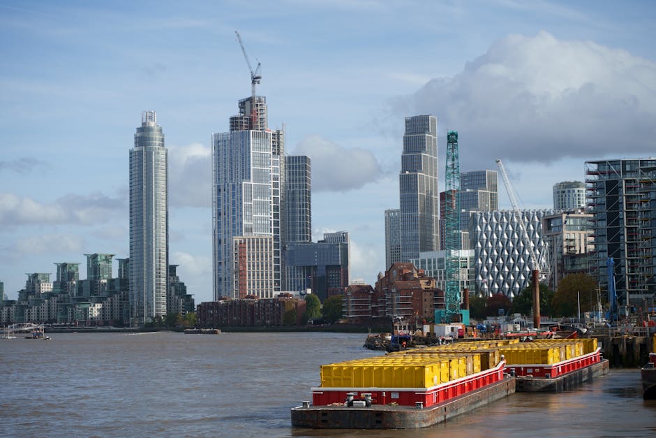 A night-time view of the London skyline featuring illuminated modern office buildings and skyscrapers along the river Thames, which reflect the city lights on the water. The buildings include tall glass structures with bright interior lighting and red aviation warning lights on the rooftops. In the foreground, there are darker, lower-rise buildings and a waterfront area with streetlights casting a warm glow on the pavement. This urban scene captures the vibrant atmosphere typical of a city engaged in commercial and residential activities, resembling the environment around the Isle of Dogs in Docklands. Such imagery is relevant to house removals, highlighting the logistics of urban moving services provided by companies like Docklands Man and Van, including loading and transport within a metropolitan setting during night hours.
