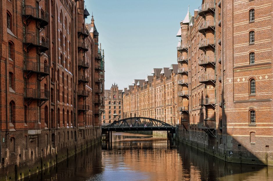 A wide view of a narrow waterway flanked by tall, historic red brick warehouse buildings with multiple small fire escape balconies on each side. The water reflects the brick facades and a small, arched metal bridge spans across the canal towards the background. The buildings have a uniform appearance with decorative brickwork and small, evenly spaced windows. The scene is outdoors in natural daylight with clear skies, and the environment resembles an area designated for house removals and home relocation services, where furniture transport and packing processes may take place near the canal. The setting suggests a location typical of docklands or portside areas, with a focus on the architectural surroundings and the logistical aspects of moving large items within this urban, waterside environment, as managed by Docklands Man and Van for small removals and narrow access solutions.