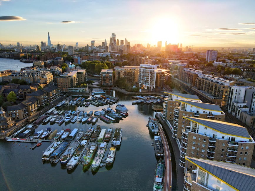 A wide view of the Docklands area with tall modern commercial and residential buildings lining both sides of a canal or river. The image shows a clear sky with some light cloud cover, and the water reflects the surrounding high-rise structures. On the left side, a dark green and black truck is partially visible, situated near the water's edge, possibly being used for home relocation or furniture transport. The buildings are constructed from glass, steel, and concrete, featuring various architectural styles including sleek curved facades and rectangular windows. The scene suggests a busy urban environment, likely associated with moving and packing services in Docklands, as depicted by the proximity of vehicles and the closeness of the buildings, which may involve narrow access solutions for household or office removals managed by Docklands Man and Van.