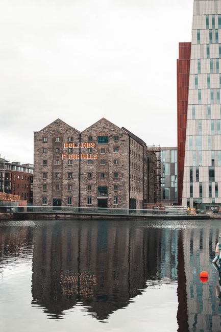 A historic brick building with orange neon signage reading 'FLOUR MILLS' is situated beside a waterway in Docklands as part of a property development, with modern glass skyscrapers nearby. The building's exterior features multiple small windows, and the water in the foreground reflects the building and the overcast sky above. To the right side of the image, a portion of a white vehicle can be seen, indicating possible loading or unloading activity. The water's edge is lined with a concrete embankment, and a small red buoy floats in the canal. Overhead, the light is diffuse due to the cloudy weather, creating a subdued atmosphere. Although no moving activity is visible, this scene illustrates an urban environment where house removals and furniture transport may occur, with potential for loading and staging in front of the building as part of a home relocation or moving process facilitated by Docklands Man and Van.