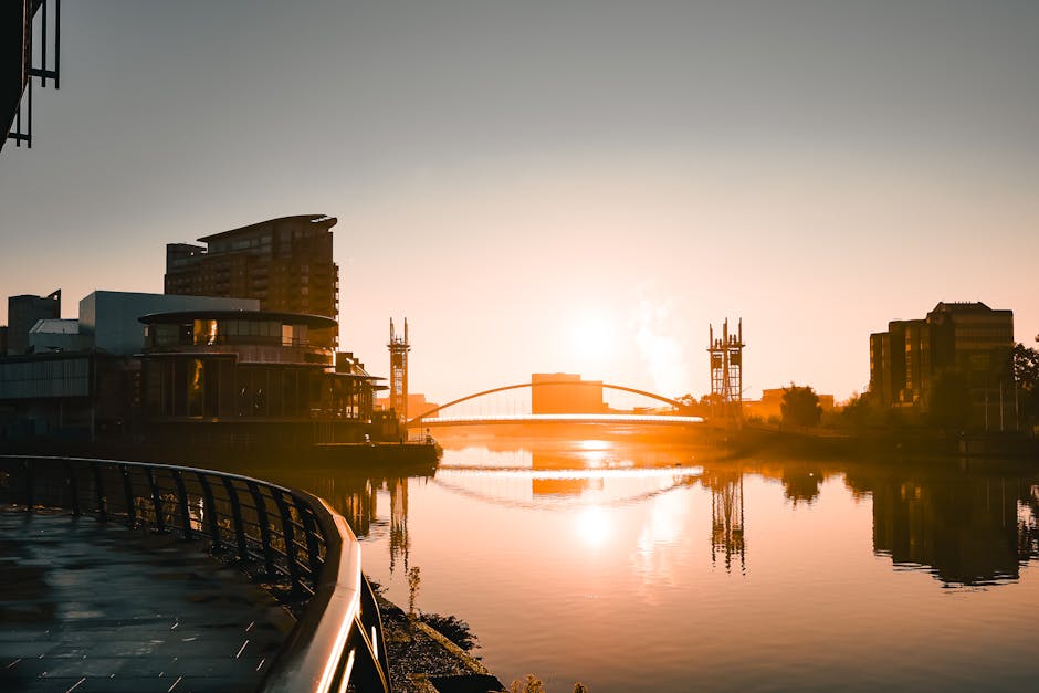 A riverside scene in Docklands during sunset, showing modern office buildings and residential structures, some with illuminated windows, reflected in calm water. In the foreground, a metal railing runs along the edge of a paved walkway, with a few cardboard boxes and packages stacked nearby, covered partially with plastic wrap and packing blankets. A hand truck or trolley is visible adjacent to the boxes, hinting at a home relocation or furniture transport process. Two large vans are parked on the pavement, with open doors and visible loading areas where furniture and boxes are being carried or transferred. The overall environment is well-lit by natural light, with the setting sun casting a warm orange glow across the scene, highlighting the busy logistics involved in house removals, packing, and moving services provided by Docklands Man and Van. The image captures key elements of the loading process, including equipment, packaging materials, and active handling of household items in preparation for transport.